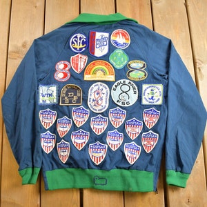 Puede incluir: Una chaqueta azul con cuello y puños verdes, cubierta de varios parches. Los parches son principalmente circulares y presentan diferentes diseños, incluidos logotipos, texto e imágenes. La chaqueta tiene una cremallera y dos bolsillos.