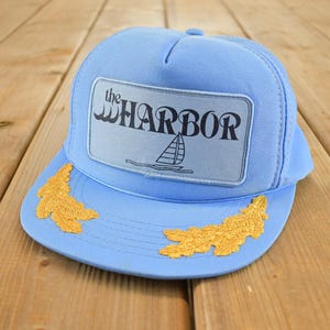 Può includere: Cappellino da camionista azzurro con una toppa rettangolare con la scritta "the HARBOR" e una grafica di una barca a vela. La visiera presenta disegni di foglie ricamate in oro. Il cappello è su una superficie di legno.