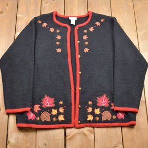 May include: Black cardigan sweater with red trim and embroidered autumn leaves in shades of brown, orange, and red. The sweater has a button closure.