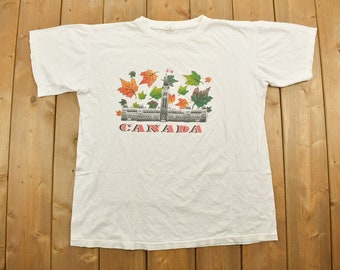 Könnte beinhalten: Weißes T-Shirt mit einem grafischen Aufdruck des kanadischen Parlamentsgebäudes mit Herbstblättern und dem darunter gedruckten Wort "CANADA".