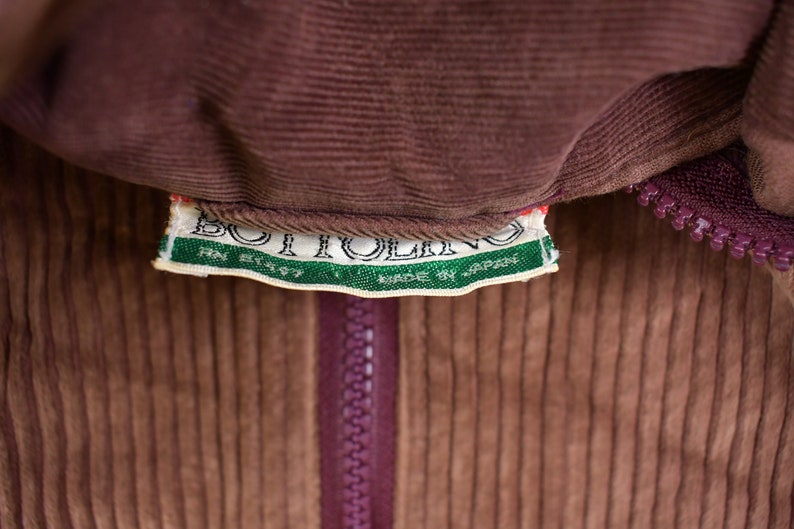 May include: A brown corduroy jacket with a zipper closure and a green and white label that reads "BOITOLLINO MADE BY JAPAN RN 81157".