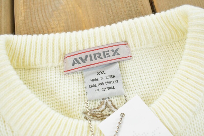 May include: Cream-colored sweater with a ribbed collar. The label reads "AVIREX" in red and white. A tag attached to the sweater displays "2XL" and "Made in Korea". The sweater has a textured knit design.