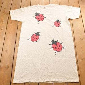 Pode incluir: Camiseta branca com um padrão repetido de joaninhas vermelhas com pintas e contornos pretos. As joaninhas estão espalhadas pela camisa. A camisa tem mangas curtas e gola redonda.