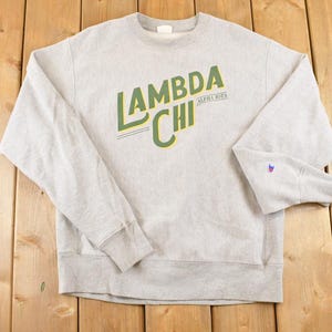 Peut inclure: Un sweat-shirt gris clair avec le texte "LAMBDA CHI ALPHA IOTA" en vert et jaune. Le sweat-shirt a des manches longues et un logo Champion sur la manche gauche.
