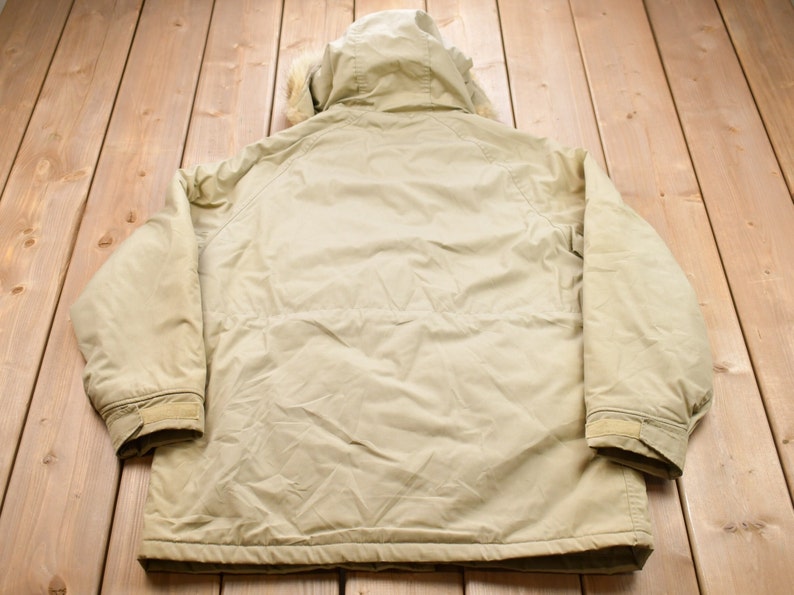 May include: A beige hooded jacket with a fur trim around the hood. The jacket has a zipper closure and two pockets on the front. The jacket is made of a water-resistant material.