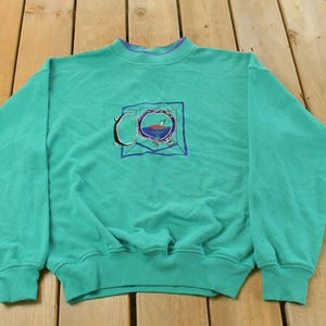 May include: Teal-colored sweatshirt with a crew neck and long sleeves. The front features an embroidered design of a circular object with a red and blue interior, framed by a blue square and a silver-colored outline. The cuffs and neckline are a darker teal.