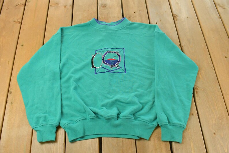 May include: Teal-colored sweatshirt with a crew neck and long sleeves. The front features an embroidered design of a circular object with a red and blue interior, framed by a blue square and a silver-colored outline. The cuffs and neckline are a darker teal.