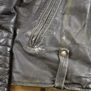 Puede incluir: Primer plano de una chaqueta de cuero negra con cremallera y cierre de presi&oacute;n. La chaqueta tiene un aspecto desgastado con costuras visibles y una superficie texturizada.