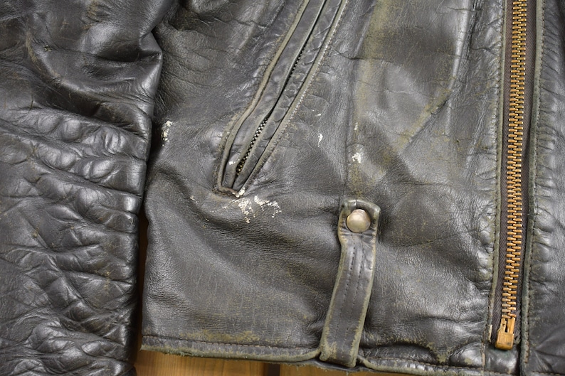Puede incluir: Primer plano de una chaqueta de cuero negra con cremallera y cierre de presi&oacute;n. La chaqueta tiene un aspecto desgastado con costuras visibles y una superficie texturizada.