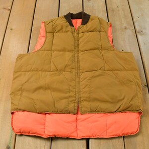 Op de afbeelding: Een omkeerbaar gewatteerd vest met een bruine buitenkant, een bruine geribbelde kraag en een fel neonoranje voering. Het vest is voorzien van een ritssluiting aan de voorzijde en een doorgestikt patroon met vierkante vakken.