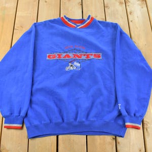 May include: A blue sweatshirt featuring the text "NEW YORK GIANTS" in red and white. The V-neck collar and cuffs have red and white trim. The design includes a football helmet graphic. The sweatshirt is a classic sports garment.