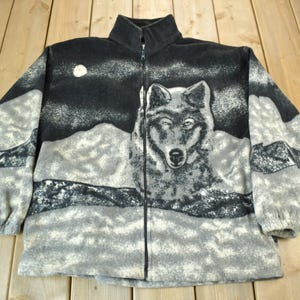 Peut inclure: Une veste en polaire noire et grise avec un imprimé représentant un loup et une scène de montagne. La veste a une fermeture éclair et un col montant.