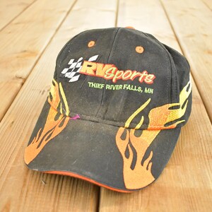 Op de afbeelding: Zwarte baseballcap met oranje en gele vlamontwerpen op de rand en zijkanten. De cap heeft de tekst "RV Sports" in rood en wit, met daaronder "Thief River Falls, MN". De cap heeft een oranje rand.