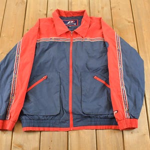 May include: A vintage navy blue and red jacket with a full zipper closure. The jacket features red accents on the shoulders, collar, and zipper, with a striped design. Two front pockets and a ribbed hem complete the design.
