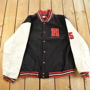 May include: Black and white varsity jacket with red detailing. The jacket has a red "H" and "5" on the front and sleeve. It features a button-up front and a red, white, and black striped trim.