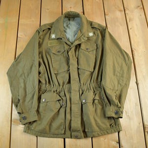 Könnte beinhalten: Olivgrüne Jacke im Militärstil mit mehreren Taschen, einer Knopfleiste und einem Kordelzug in der Taille. Die Jacke hat einen Kragen und Schulterklappen mit sternförmigen Abzeichen. Die Jacke liegt auf einer Holzoberfläche.