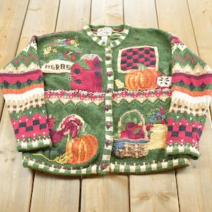 Peut inclure: Un cardigan vert à manches longues boutonné avec un motif tricoté et des broderies représentant des citrouilles, un panier de produits et le mot HERBS. Les manches et les bordures présentent un motif géométrique rouge, blanc et beige.