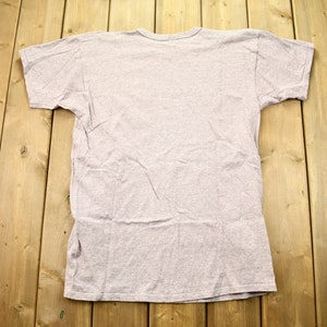 May include: A plain, light gray t-shirt with short sleeves. The shirt is laid flat on a wooden surface.