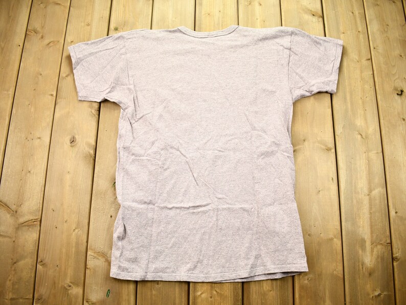 May include: A plain, light gray t-shirt with short sleeves. The shirt is laid flat on a wooden surface.
