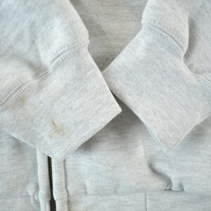 Puede incluir: Primer plano de una sudadera con capucha gris claro con cremallera. Se ven los pu&ntilde;os de las mangas, que muestran un dise&ntilde;o de punto acanalado. Hay algunas manchas en el pu&ntilde;o izquierdo. La cremallera es parcialmente visible.