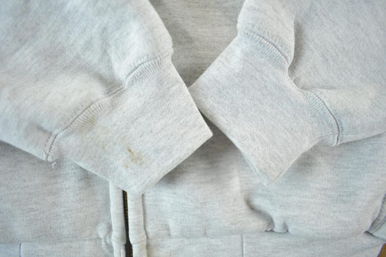 Puede incluir: Primer plano de una sudadera con capucha gris claro con cremallera. Se ven los pu&ntilde;os de las mangas, que muestran un dise&ntilde;o de punto acanalado. Hay algunas manchas en el pu&ntilde;o izquierdo. La cremallera es parcialmente visible.