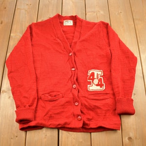 Pode incluir: Um cardigan vermelho de lã com um remendo de feltro branco na frente que diz "44".