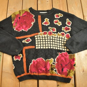 May include: Black knitted sweater with a floral pattern in shades of red, orange, and green. The sweater has a crew neck and long sleeves.