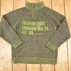 May include: Olive green knit sweater with a half-zip collar. The front features the text "PREMIUM GRADE CONFORM REG. P.Z. G2 55" in green. The sweater has ribbed cuffs and hem.