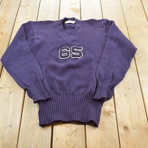 Könnte beinhalten: Ein dunkelvioletter Langarmpullover mit V-Ausschnitt und der Zahl 65, die in Weiß mit dunkelviolettem Umriss auf der Brust eingestickt ist. Das Kleidungsstück hat gerippte Bündchen und einen breiten gerippten Saum für eine strukturierte Passform.