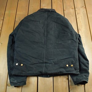 Puede incluir: Chaqueta de trabajo negra con cuello marr&oacute;n y botones dorados. La chaqueta est&aacute; hecha de un material duradero y tiene un dise&ntilde;o cl&aacute;sico. La parte trasera de la chaqueta es visible, mostrando las costuras y los detalles de construcci&oacute;n.