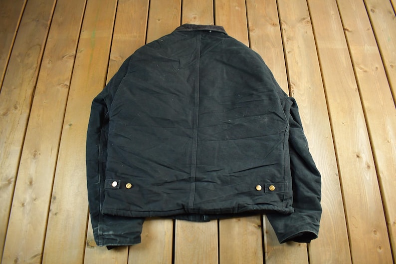 Puede incluir: Chaqueta de trabajo negra con cuello marr&oacute;n y botones dorados. La chaqueta est&aacute; hecha de un material duradero y tiene un dise&ntilde;o cl&aacute;sico. La parte trasera de la chaqueta es visible, mostrando las costuras y los detalles de construcci&oacute;n.