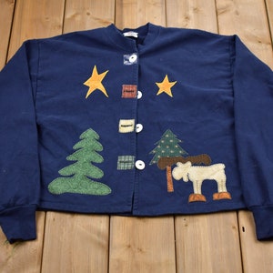Peut inclure: Une veste boutonnée à manches longues bleu marine avec un motif en patchwork représentant un élan, des arbres et des étoiles.