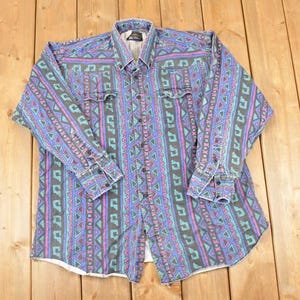 May include: A long-sleeved button-down shirt with a colorful geometric pattern. The shirt features a mix of blue, purple, and teal vertical stripes with repeating shapes. It has two chest pockets and buttoned cuffs.