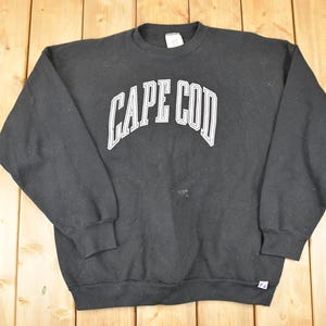 May include: Black crew neck sweatshirt with the text "CAPE COD" in white, arched across the chest. The sweatshirt is made of a soft material and has long sleeves. A small stain is visible on the front.