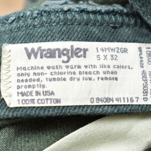 Op de afbeelding: Een wit waslabel bevestigd aan een donkergroen kledingstuk. Het label vermeldt "Wrangler", "14MWZGR", "5 X 32", "Machinewas warm met soortgelijke kleuren, alleen chloorvrij bleekmiddel gebruiken indien nodig, drogen in de droger op lage temperatuur, snel verwijderen.", "MADE IN USA", "100% katoen", en "0 84084 411167".
