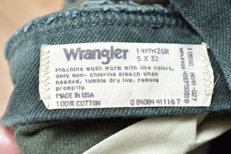 Op de afbeelding: Een wit waslabel bevestigd aan een donkergroen kledingstuk. Het label vermeldt "Wrangler", "14MWZGR", "5 X 32", "Machinewas warm met soortgelijke kleuren, alleen chloorvrij bleekmiddel gebruiken indien nodig, drogen in de droger op lage temperatuur, snel verwijderen.", "MADE IN USA", "100% katoen", en "0 84084 411167".