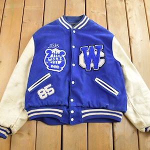 May include: A blue and white varsity jacket with white leather sleeves. The jacket has a large "W" on the right side and a smaller patch with a lyre and the text "WCHS Band" on the left side. The number "85" is on the left side of the jacket. The jacket has a white zipper and white stripes on the cuffs and waistband.