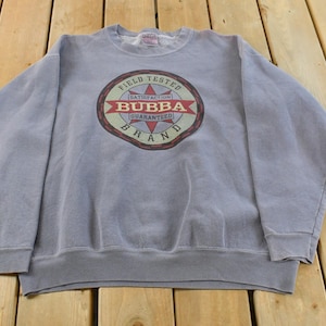 Könnte beinhalten: Hellblaues Sweatshirt mit Rundhalsausschnitt und langen Ärmeln. Auf der Vorderseite befindet sich ein kreisförmiges Grafikdesign mit den Worten "FIELD TESTED", "BUBBA BRAND" und "SATISFACTION GUARANTEED" in Rot, Braun und Weiß.