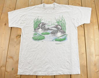 May include: A light gray short-sleeve t-shirt featuring a graphic print of two loons swimming in a lake with green reeds and lily pads. The word MINNESOTA is printed in small black letters below the water illustration on the front of the shirt.