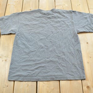 May include: A gray, short-sleeved t-shirt, viewed from the back, lies on a light-colored wooden surface. The shirt has a crew neck and appears to be made of a soft, slightly wrinkled fabric. The background is a light-colored wooden surface.