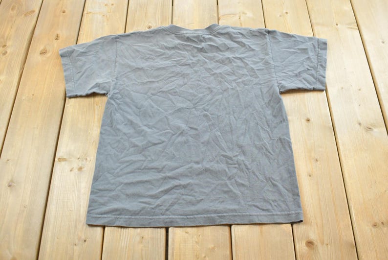 May include: A gray, short-sleeved t-shirt, viewed from the back, lies on a light-colored wooden surface. The shirt has a crew neck and appears to be made of a soft, slightly wrinkled fabric. The background is a light-colored wooden surface.
