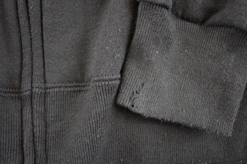 May include: Close-up of a dark gray zip-up hoodie. The image shows the fabric texture, seams, and a torn cuff. The hoodie appears to be well-worn, with visible wear and tear on the sleeve.