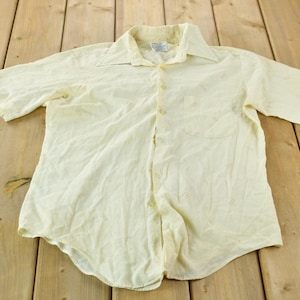 Puede incluir: Una camisa de manga corta de color amarillo pálido con botones y cuello clásico.