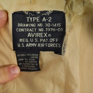 May include: Close-up of a black label from an A-2 jacket, with text including "TYPE A-2", "AVIREX", and "U.S. ARMY AIR FORCES". The label also lists the materials: 100% genuine leather, 50% nylon, and 50% acetate.