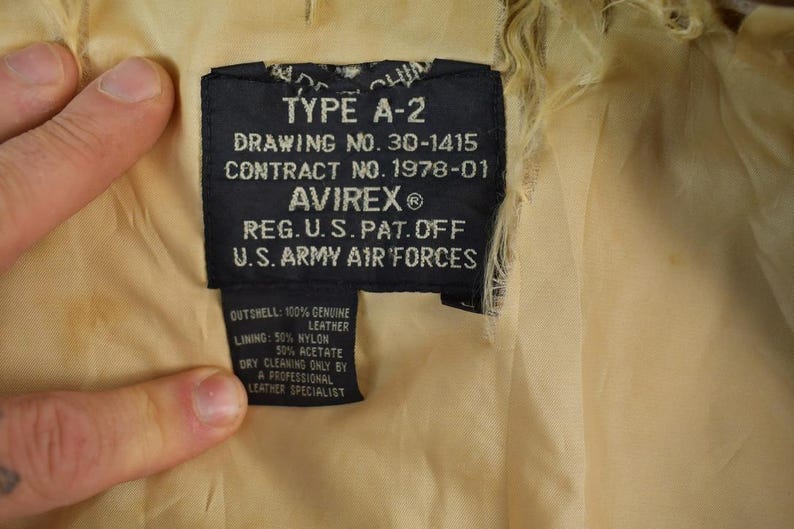 May include: Close-up of a black label from an A-2 jacket, with text including "TYPE A-2", "AVIREX", and "U.S. ARMY AIR FORCES". The label also lists the materials: 100% genuine leather, 50% nylon, and 50% acetate.