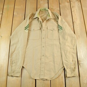 Op de afbeelding: Beige overhemd met knopen, twee borstzakken en groene strepen op de schouders. Het shirt met lange mouwen heeft een kraag en manchetten met knopen. Het shirt ligt plat op een houten oppervlak.