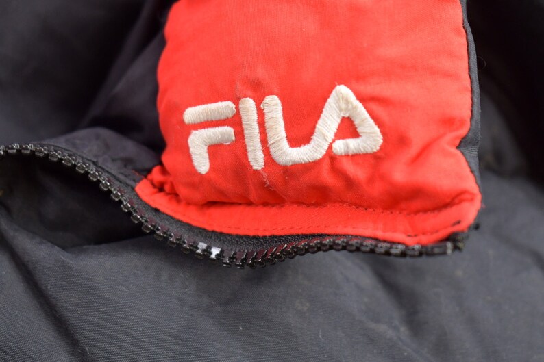 May include: Close-up of a black and red jacket with a white embroidered FILA logo on the red section.