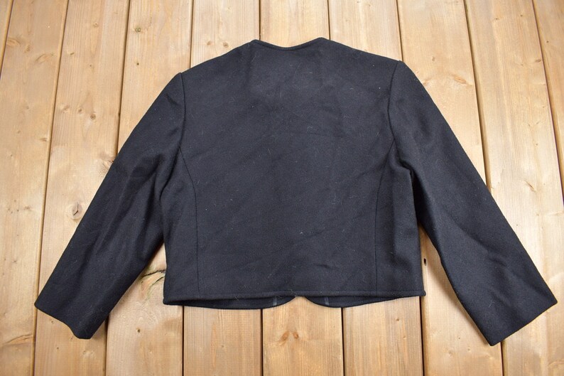 K&ouml;nnte beinhalten: Schwarzer Blazer mit langen &Auml;rmeln, von hinten betrachtet. Die Jacke hat ein klassisches Design mit rundem Ausschnitt und taillierter Passform. Der Stoff ist dunkel und einfarbig, geeignet f&uuml;r formelle oder halbformelle Anl&auml;sse.