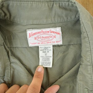 Peut inclure: Gros plan sur un v&ecirc;tement vert olive d&eacute;lav&eacute; avec un col et une &eacute;tiquette. L'&eacute;tiquette indique "A GENUINE FILSON GARMENT" et "CC-FILSON CO. SEATTLE". Le v&ecirc;tement est en 100% coton et est de style 58, taille 44.