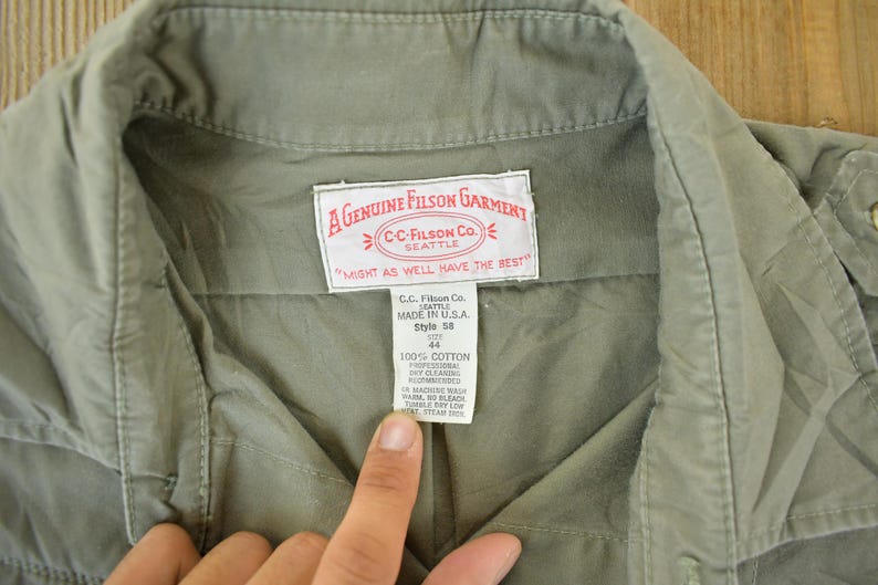 Peut inclure: Gros plan sur un v&ecirc;tement vert olive d&eacute;lav&eacute; avec un col et une &eacute;tiquette. L'&eacute;tiquette indique "A GENUINE FILSON GARMENT" et "CC-FILSON CO. SEATTLE". Le v&ecirc;tement est en 100% coton et est de style 58, taille 44.